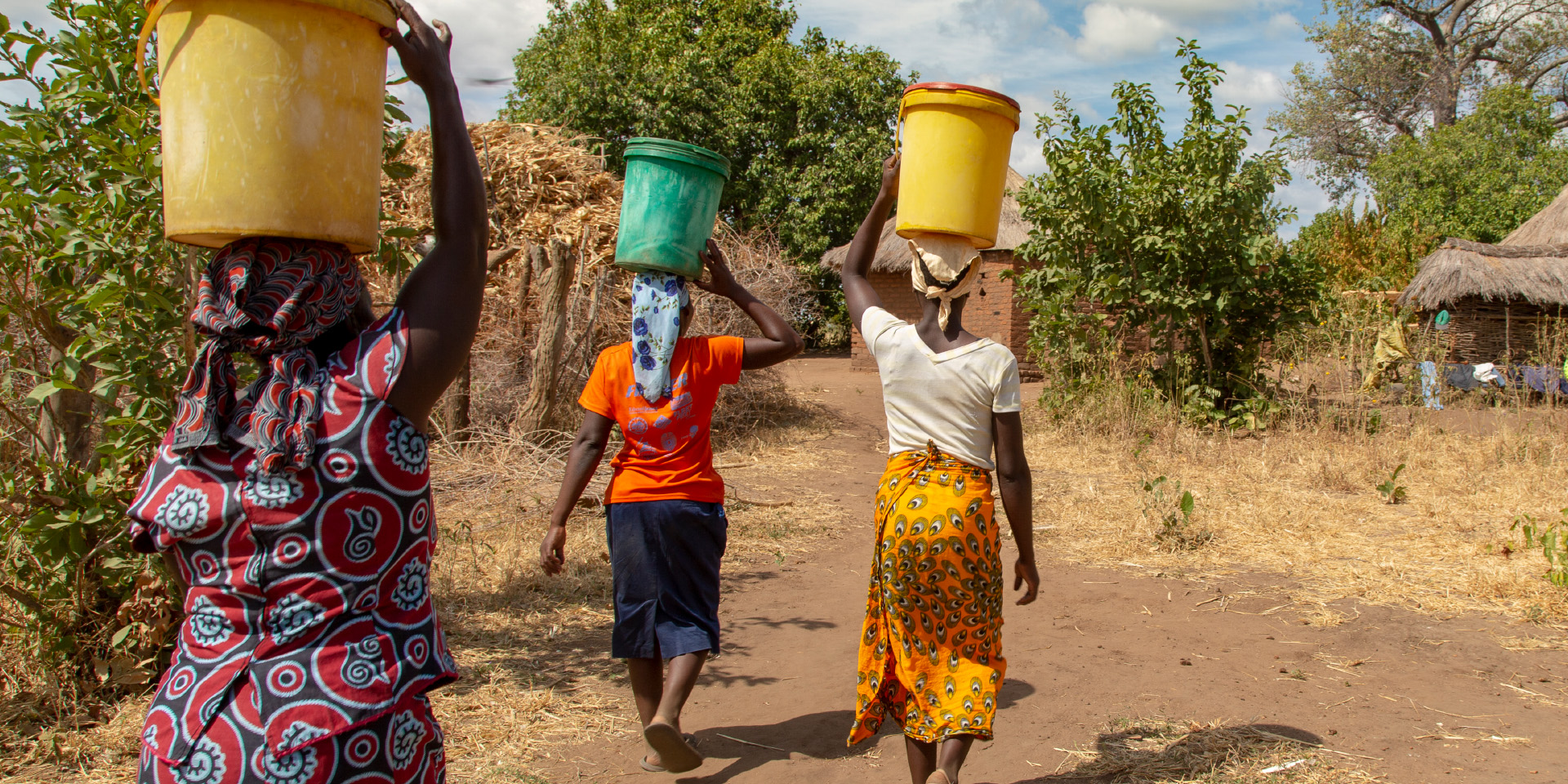 A shot of three women from behind, following a dried-up path. They have dark skin tones and are wearing skirts and headscarves. Each woman is balancing a large bucket on her head and stabilizing it with one hand. In the distance, trees and a small straw-roofed house can be seen.